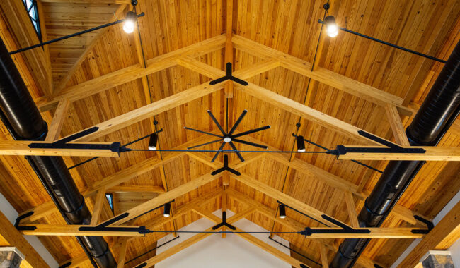 Wooden vaulted ceiling with exposed beams, black metal supports, ductwork, and a large ceiling fan in the center. Multiple light fixtures are mounted along the beams, and natural light enters through small skylights.