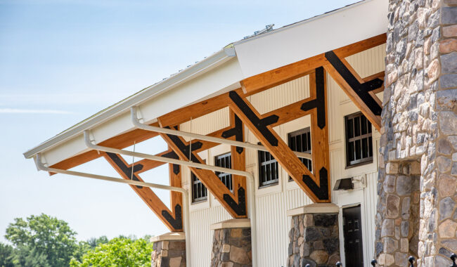 A close-up view of a building with stone pillars, white siding, and exposed wooden beams supporting the roof, photographed on a sunny day with trees visible in the background.