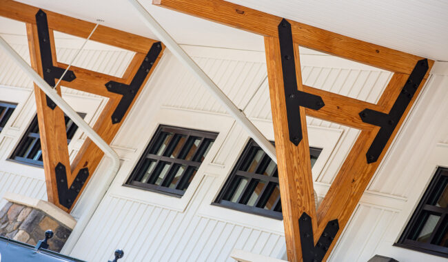 Large, decorative wooden support beams with black metal brackets are attached to a white building with rectangular windows, stone accents at the base, and a white ceiling above.