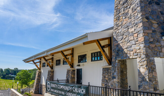 A stone and white building with a sign reading Trinity Community Church, Dean Lancaster Park—Sundays 10 AM, set against a blue sky and green landscape.