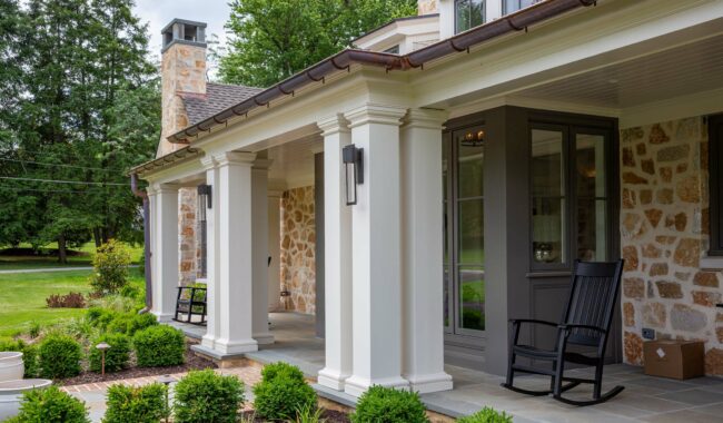 A stone and brick house with a covered porch featuring white columns, two black rocking chairs, a manicured lawn, shrubs, and trees in the background.