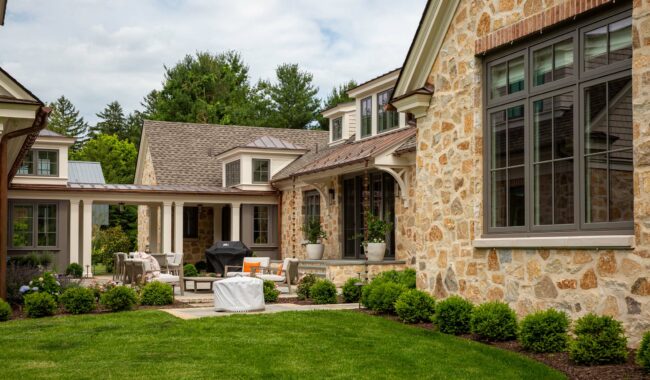 A stone house with large windows and a neatly manicured lawn. There is a covered patio area with outdoor furniture and a barbecue grill, surrounded by green shrubs and trees in the background.