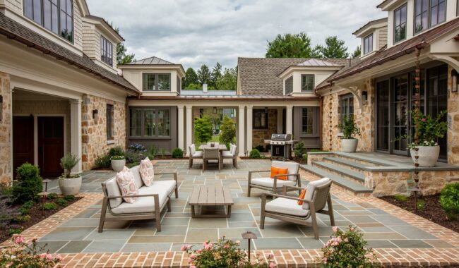 Spacious patio with cushioned chairs, a coffee table, and potted plants, surrounded by a stone and brick walkway. Outdoor dining area and grill are visible, set between two sections of a large, elegant house.