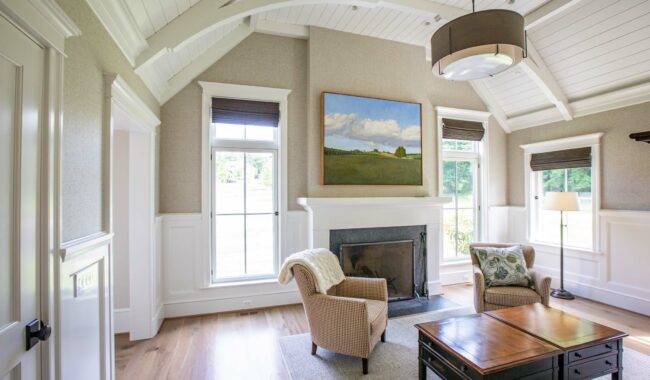 Bright living room with vaulted white ceiling, light walls, and large windows. Features a fireplace with a landscape painting above, two armchairs, a wooden coffee table, and a floor lamp. Natural light fills the space.