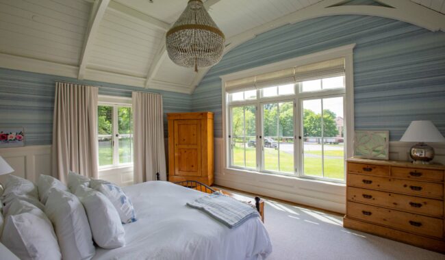 A bright, airy bedroom with a white vaulted ceiling, large windows, a bed with white linens and pillows, a wooden dresser, a wardrobe, and a chandelier. Sunlight streams in, highlighting the serene, cozy atmosphere.