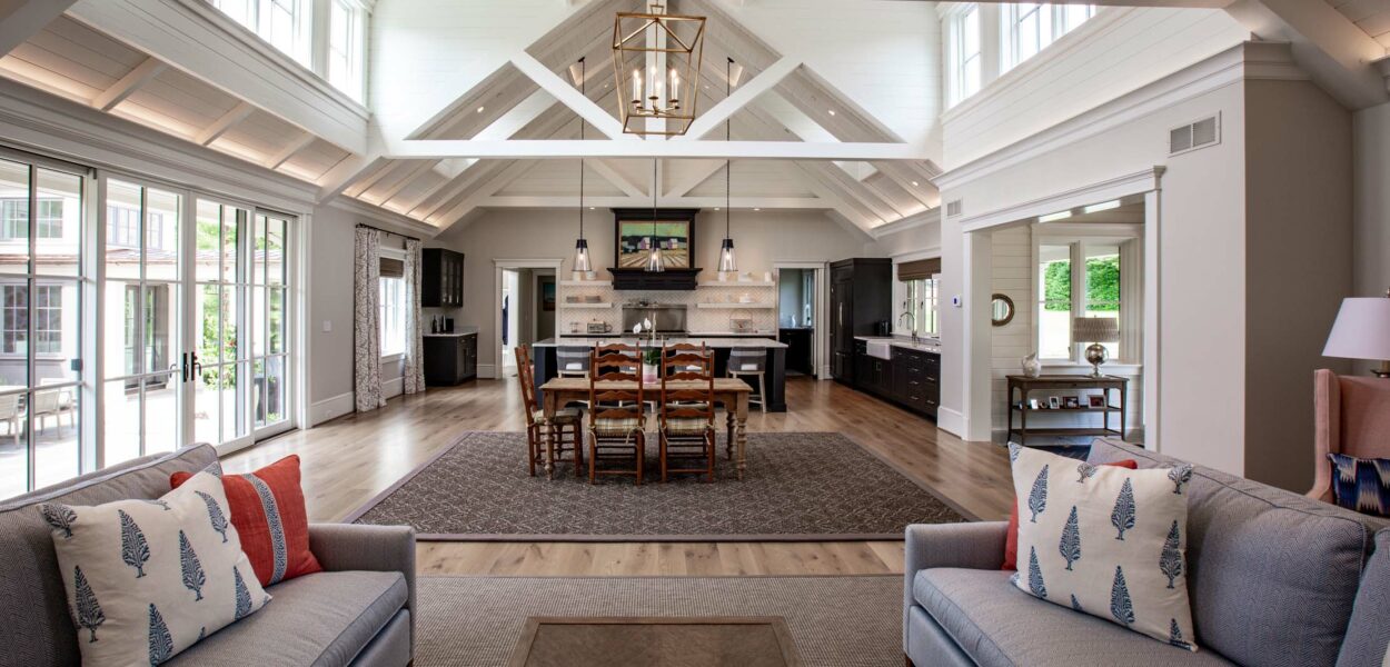 Bright, spacious open-concept living area with vaulted ceilings, exposed beams, two facing sofas, a large dining table, kitchen island, and natural light streaming through large windows and glass doors.