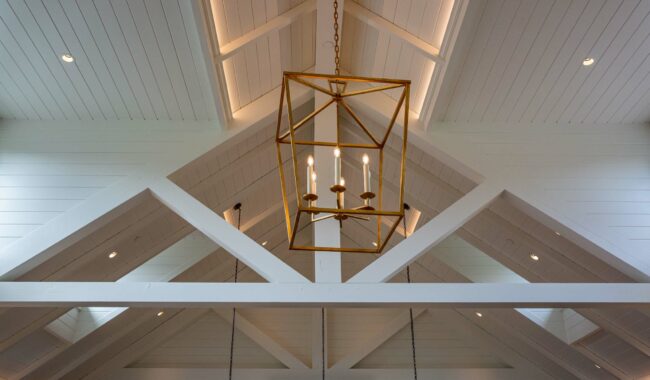 A modern, vaulted ceiling with exposed white beams and a large, gold geometric chandelier with four candle-style lights hanging in the center. Recessed lighting highlights the ceiling architecture.
