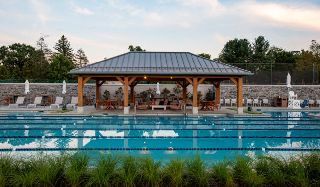 A swimming pool with clear blue water is in the foreground, bordered by green plants. Behind the pool, there is a covered seating area with wooden furniture and umbrellas, set against a stone wall and surrounded by trees.