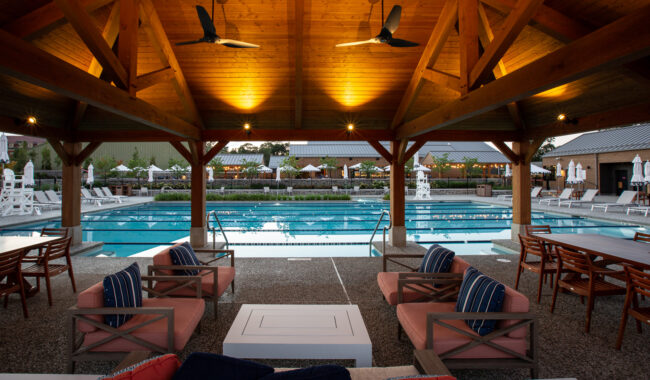 Under a wooden pavilion with ceiling fans, cushioned lounge seating and tables face a large outdoor pool. The well-lit area includes pool chairs and umbrellas in the background, creating a relaxing atmosphere.
