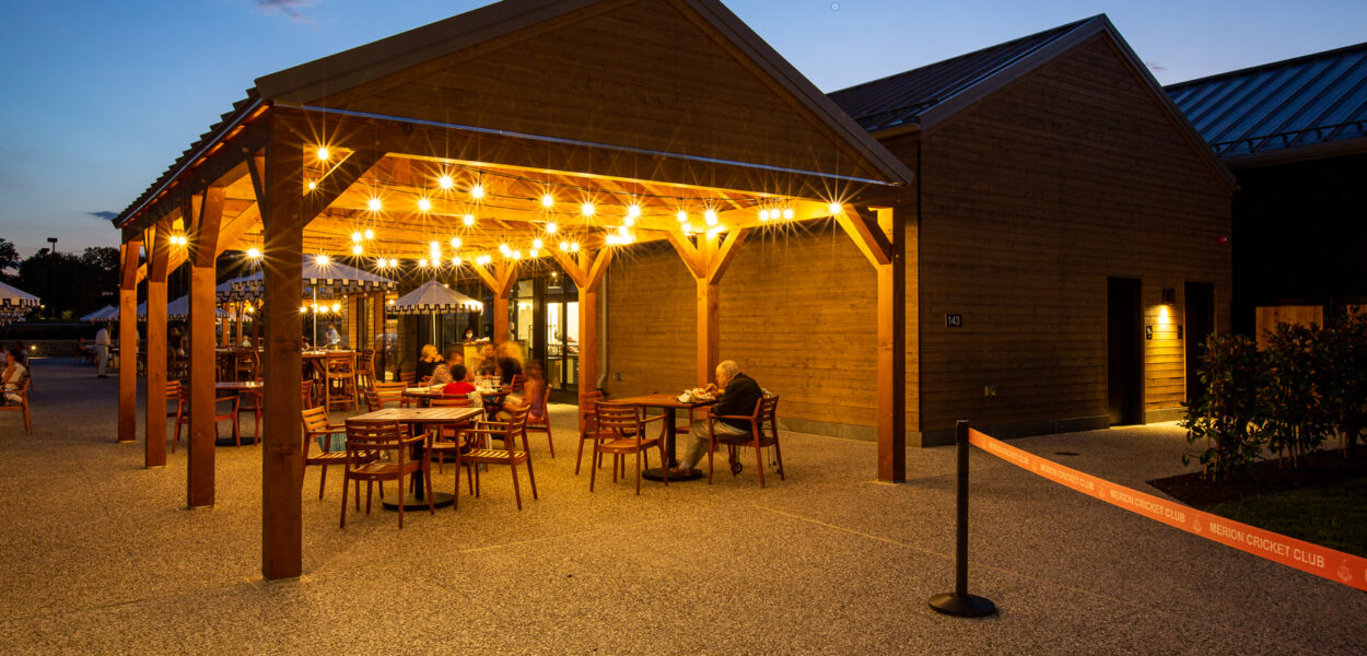 A covered outdoor patio at dusk, with string lights hanging from the wooden beams. Several people sit at tables under the pavilion, while a nearby building is softly lit. A red barrier rope is in the foreground.