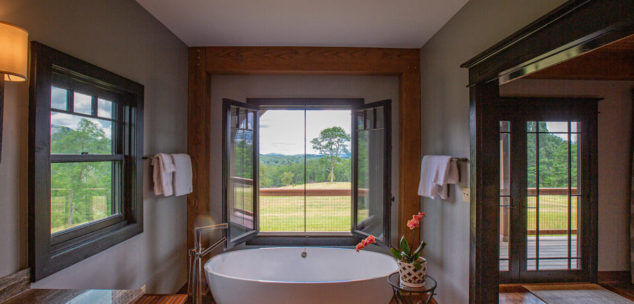A modern bathroom with a freestanding white bathtub by large open windows overlooking a green landscape, wooden floors, gray walls, and a small table with a potted plant beside the tub.