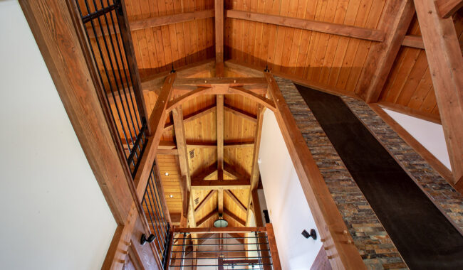 View looking up at a vaulted ceiling with exposed wooden beams and trusses in a rustic interior. Metal railings and a stone accent wall are also visible, contributing to the architectural design.