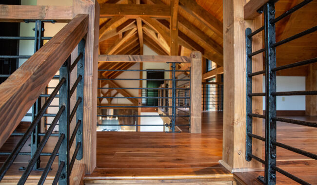 Sunlit loft area with exposed wooden beams, polished hardwood floors, and a railing with wooden posts and black metal bars, creating a warm, rustic interior space.