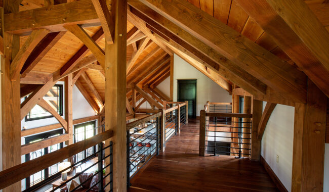 A loft with exposed wooden beams, slanted ceiling, hardwood floors, metal railing, and natural light coming through large windows, creating a warm and rustic atmosphere.