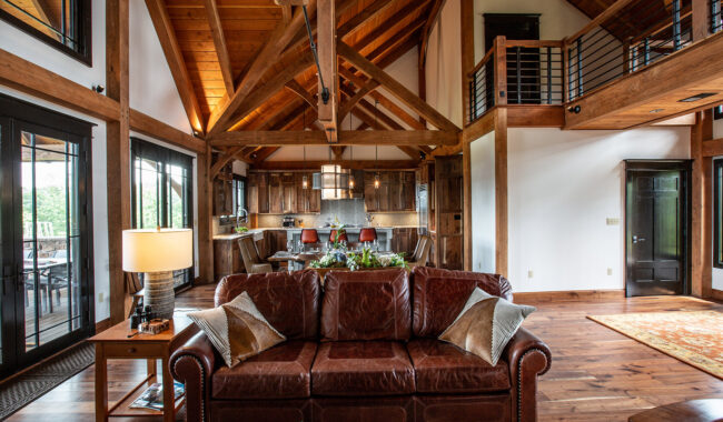 A rustic living room with exposed wooden beams, a brown leather sofa, high ceilings, and an open kitchen in the background, featuring wooden cabinets and a loft area above. Natural light fills the space.