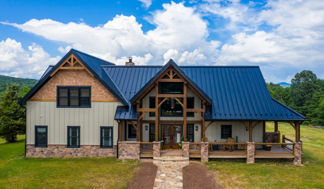 A modern farmhouse with a metal roof, timber framing, stone accents, and a wraparound porch, set among green lawns and trees under a partly cloudy blue sky.