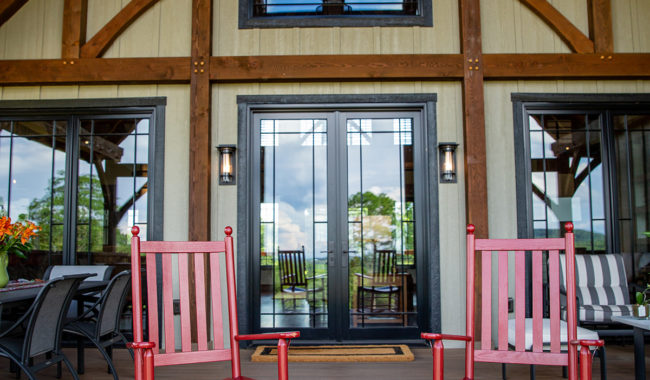 Two red rocking chairs sit on a wooden porch in front of glass double doors, with a modern rustic house facade, exposed wooden beams, and outdoor seating area visible on either side.