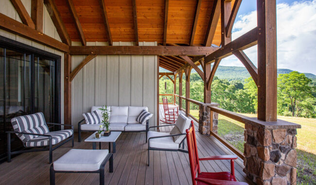 A covered wooden porch with cushioned seating, striped armchairs, red rocking chairs, and a view of green trees and distant mountains under a partly cloudy sky.