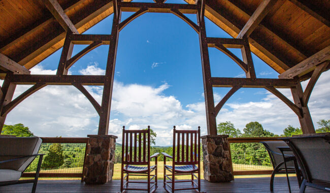 Two wooden rocking chairs sit on a covered patio with a timber-frame roof, facing out toward a scenic view of trees and a bright blue sky with clouds. The scene is inviting and peaceful.