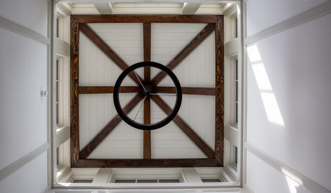 View looking up at a square coffered ceiling with wooden beams forming a star pattern and a black metal circular chandelier in the center, with sunlight streaming through side windows.