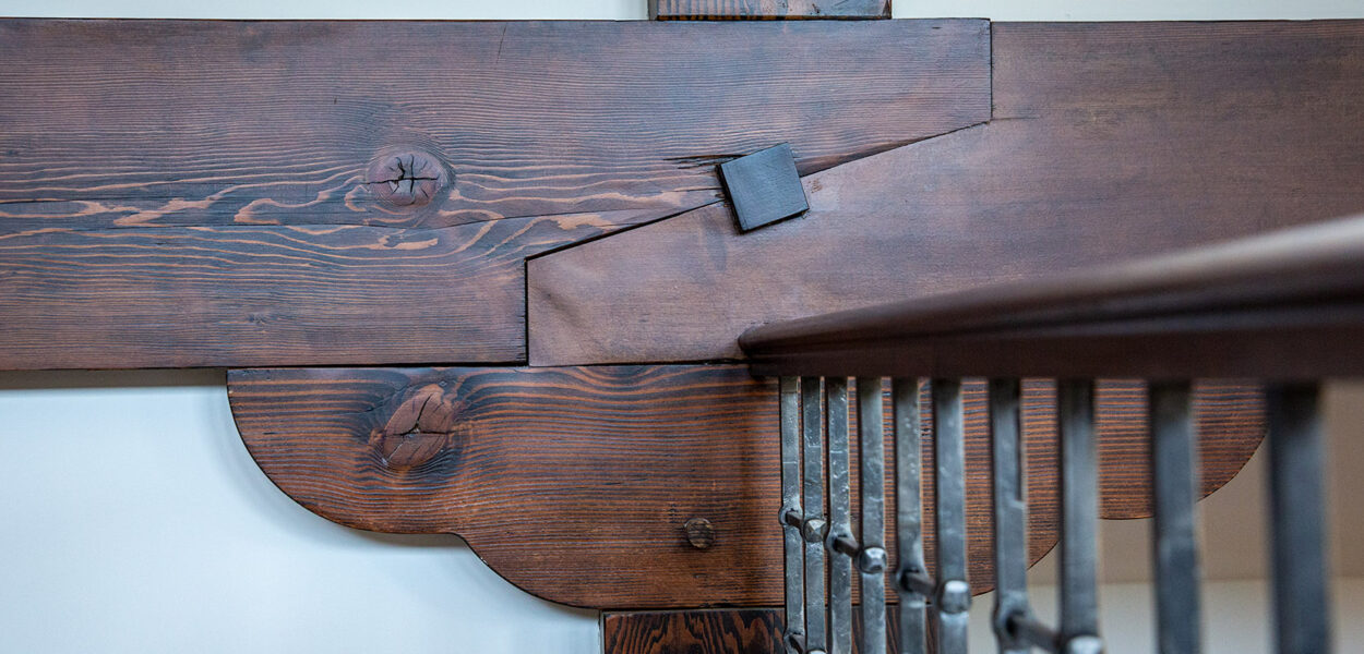 Close-up of a wooden beam with decorative detailing and a metal connector, next to a metal railing with vertical bars. The wood has a rich, dark finish and visible grain patterns.