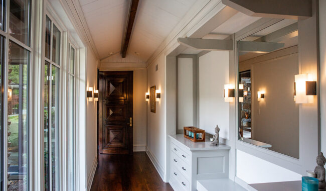 A bright hallway with large windows on the left, wooden floor, white walls, a dark wooden door at the end, built-in white drawers and mirrors on the right, and modern wall sconces providing light.
