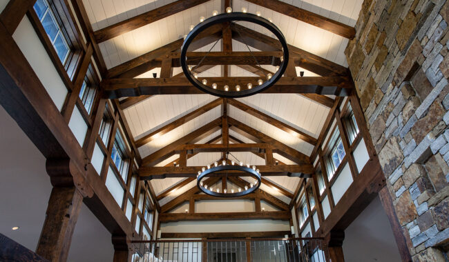 A vaulted ceiling with exposed wooden beams, circular chandeliers, white paneling, tall windows, and a stone accent wall in a rustic, modern interior space.