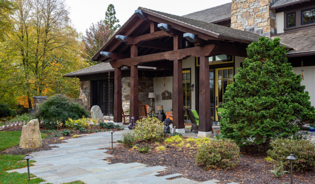 A rustic house with stone and wood exterior, a covered front porch, and landscaped garden featuring shrubs, trees, and a stone pathway leading to the entrance.
