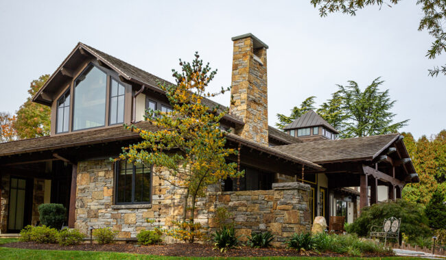 A two-story house with stone and wood exterior, large windows, and a prominent stone chimney, surrounded by green lawn, trees, and shrubs on a clear day.