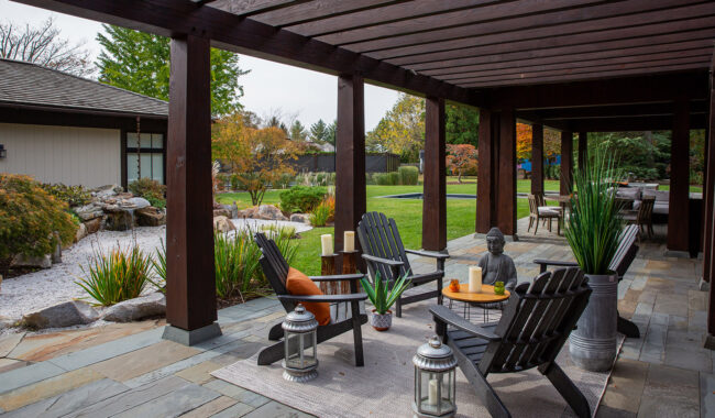 A covered patio with dark wooden beams features black Adirondack chairs, potted plants, candles, lanterns, and a small Buddha statue. The patio overlooks a landscaped garden with rocks, greenery, and a lawn.