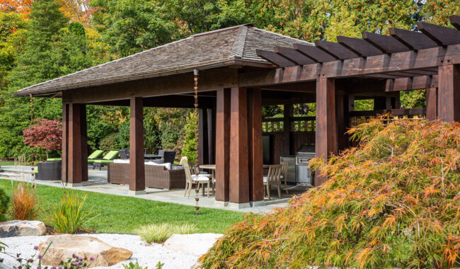 A wooden pergola and covered patio with outdoor seating and a built-in grill, surrounded by lush greenery and landscaped garden.