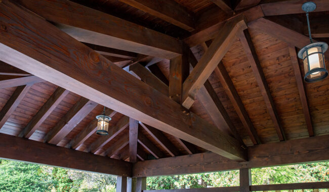 Wooden pergola ceiling with exposed beams and trusses, featuring two hanging lantern-style lights. Trees and greenery are visible outside through the open sides.