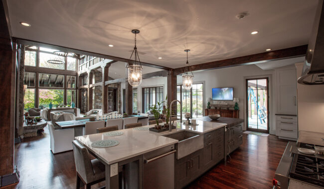 Spacious modern kitchen with a large island, double sinks, and barstools, opening to a living area with large windows, natural light, plants, and a view of a garden and patio outside.