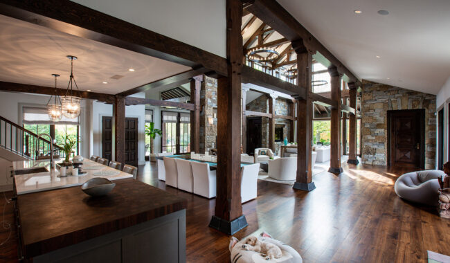 Spacious, open-concept living area with dark wood beams, stone walls, and large windows. Modern kitchen, long dining table, cozy lounge, and a dog resting on a bed on the wooden floor. Natural light fills the room.