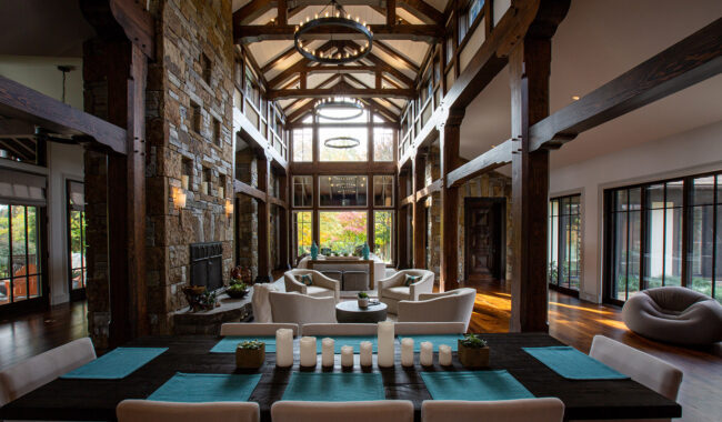 A spacious, modern rustic living and dining area with exposed wooden beams, large windows, a stone fireplace, neutral-colored furniture, and a dark wooden dining table set with candles and blue placemats.