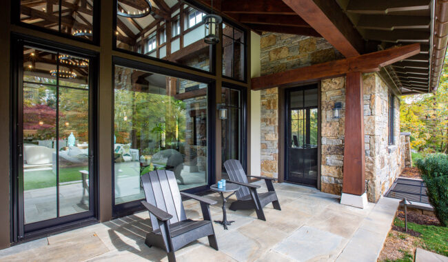 A modern patio with two black wooden chairs facing a glass wall, showing a view into a stylish living room. The patio is paved with stone and attached to a house with stone and wood exterior. Trees are visible in the background.