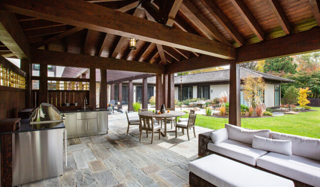 Covered patio area with wooden beams, outdoor kitchen, dining table with chairs, and white cushioned sofa. Stone flooring leads to a green lawn and landscaped garden with shrubs and trees.