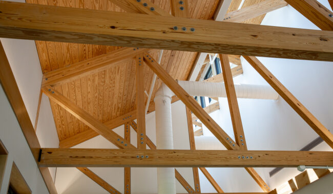 Exposed wooden beams and trusses support a sloped ceiling with natural wood paneling. White metal ducts and pipes are visible, and sunlight streams in from windows, creating a bright, airy interior.