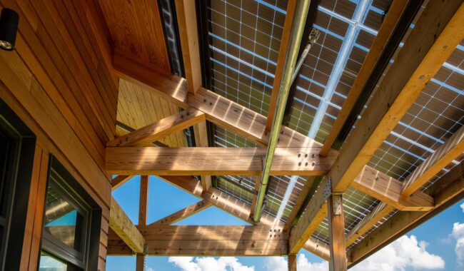 View from below of a wooden roof structure with exposed beams and integrated solar panels, set against a blue sky with scattered clouds. Sunlight streams through the semi-transparent solar panels.