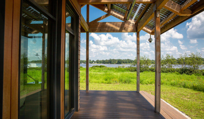 A wooden deck with a slatted roof extends from a modern building, overlooking a grassy field and a lake under a blue sky with scattered clouds.