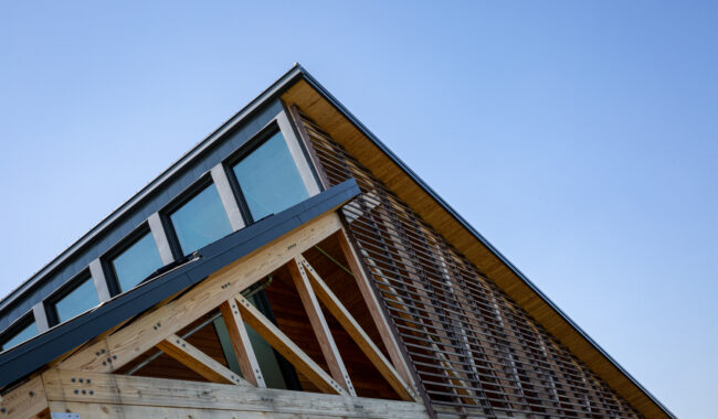 A modern building with large windows and a slanted roof, featuring exposed wooden beams and slatted wood siding, set against a clear blue sky.