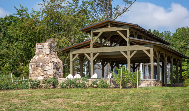 A wooden pavilion with a gabled roof stands beside a large stone outdoor fireplace. Four white Adirondack chairs are arranged under the pavilion, surrounded by grass and trees on a sunny day.