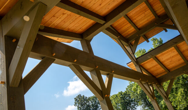 View from below of a wooden pavilion structure with exposed beams and a light wood roof, set against a blue sky and surrounded by green trees. Sunlight highlights the wood grain and details of the construction.