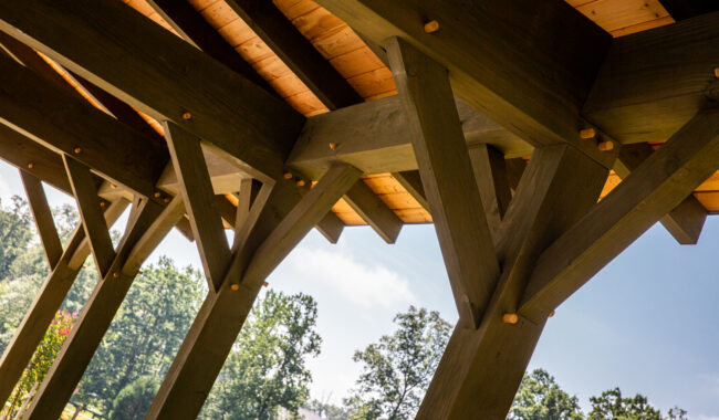 Close-up view of sturdy wooden beams and supports under a pavilion roof, with sunlight illuminating the wood and a background of green trees and blue sky.