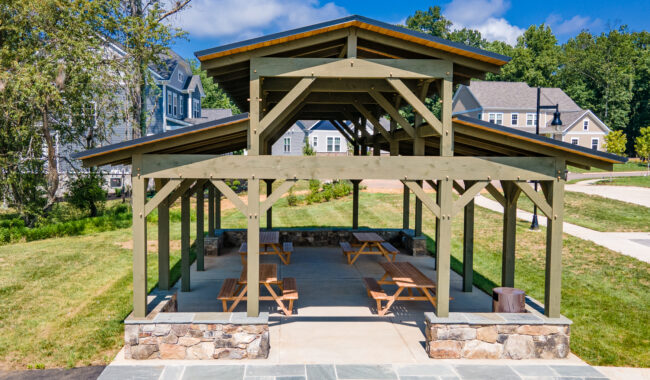 A wooden pavilion with a stone base and picnic tables underneath sits on a grassy area near houses, under a blue sky with scattered clouds.