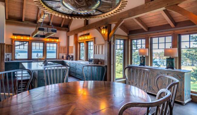 A rustic kitchen and dining area with wood beams, large windows offering a scenic lake view, a round wooden dining table with chairs, blue cabinetry, and lantern-style pendant lighting.