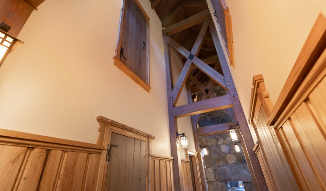 A hallway with wooden paneling, exposed beams, and stone wall accents, featuring several wooden doors and warm lantern-style wall lights illuminating the space.