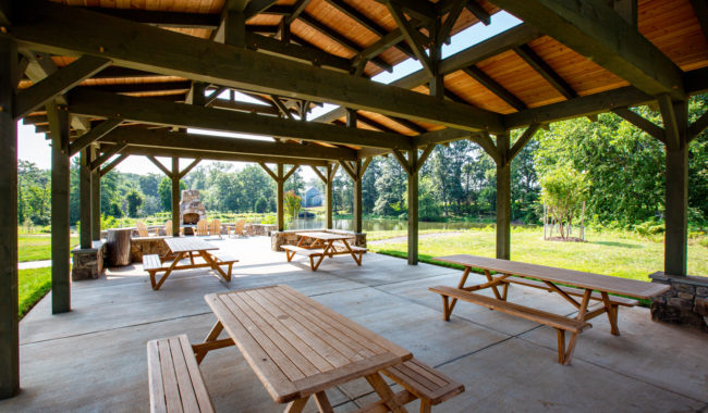 Covered outdoor pavilion with wooden tables and benches on a concrete floor, surrounded by green grass and trees, and a stone fireplace in the background near a pond.