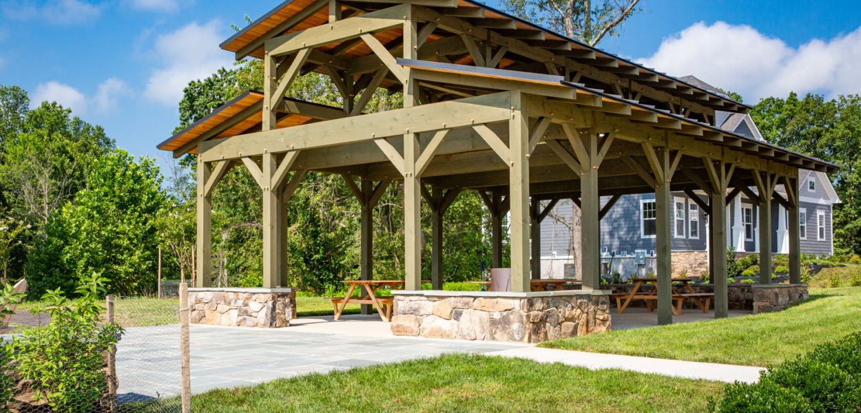A wooden pavilion with a stone base and picnic tables underneath sits on a concrete pad in a grassy yard, with trees and a house visible in the background under a blue sky with scattered clouds.