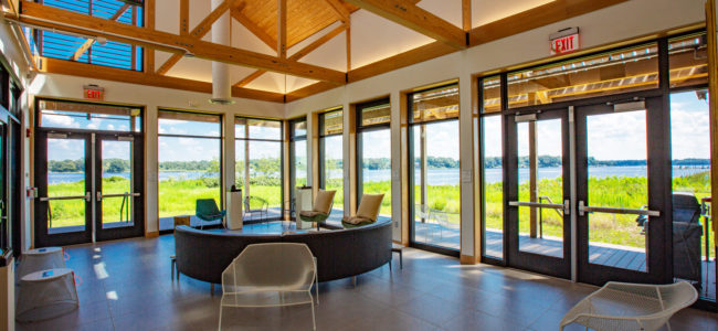 A bright, open room with large windows, wooden beams, and modern seating, offering scenic views of a lush green landscape and water outside under a blue sky.